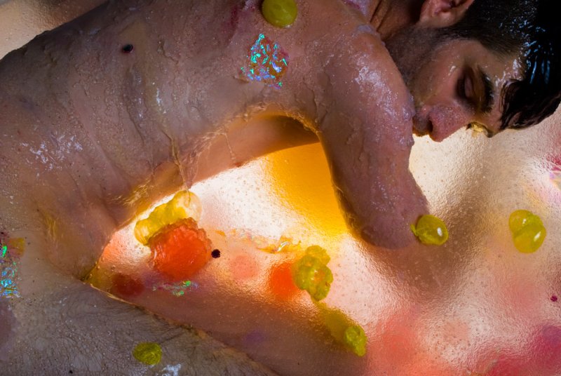Man submerged in translucent gel with colorful, iridescent shapes and yellow blobs, lit from below in warm tones.