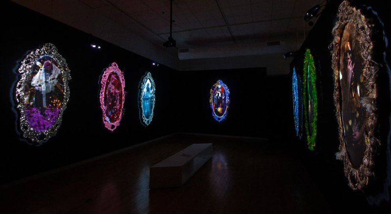 Dark room with five illuminated, ornate, oval mirror-like projections on walls, displaying colorful, abstract figures.