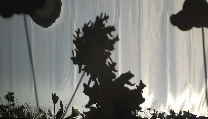 Shadow puppet show with a large tree-like figure, bears, and other animals silhouetted against a bright white screen.