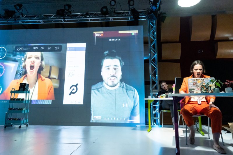 Installation view with a large screen projecting two video portraits and data. A woman in an orange suit sits at a desk, mimicking the expression of one of the video portraits. Another person sits at a desk in the background.