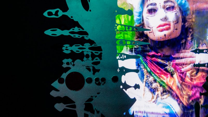 Abstract artwork featuring a figure with surgical markings, overlaid with teal geometric shapes on a black background. The figure is draped in colorful fabrics with a distorted, digital effect.