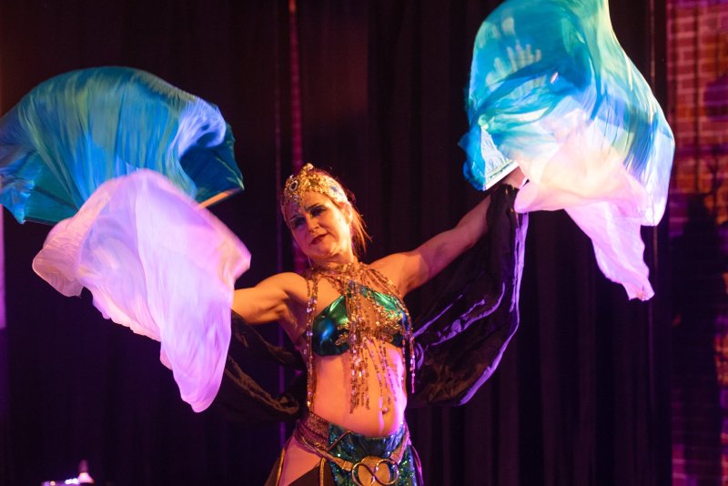 Belly dancer in a gold and teal costume with a crown, holding flowing blue and purple silk fans against a dark background.