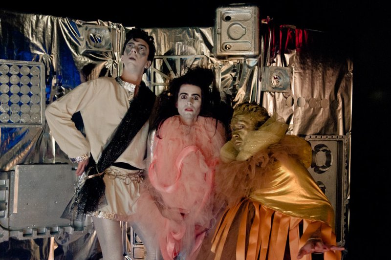 Three performers in elaborate, futuristic costumes against a metallic, textured backdrop.