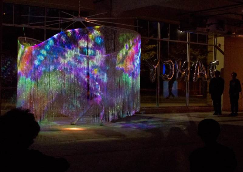Light installation with shimmering beaded curtain projecting colorful abstract patterns, viewed by silhouetted figures.