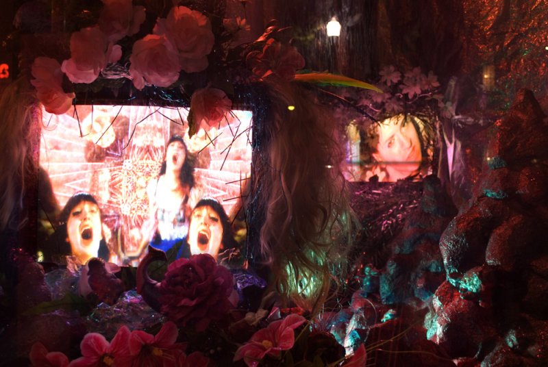 Multimedia installation with screens showing screaming faces, surrounded by pink flowers, dark foliage, and glowing lights.