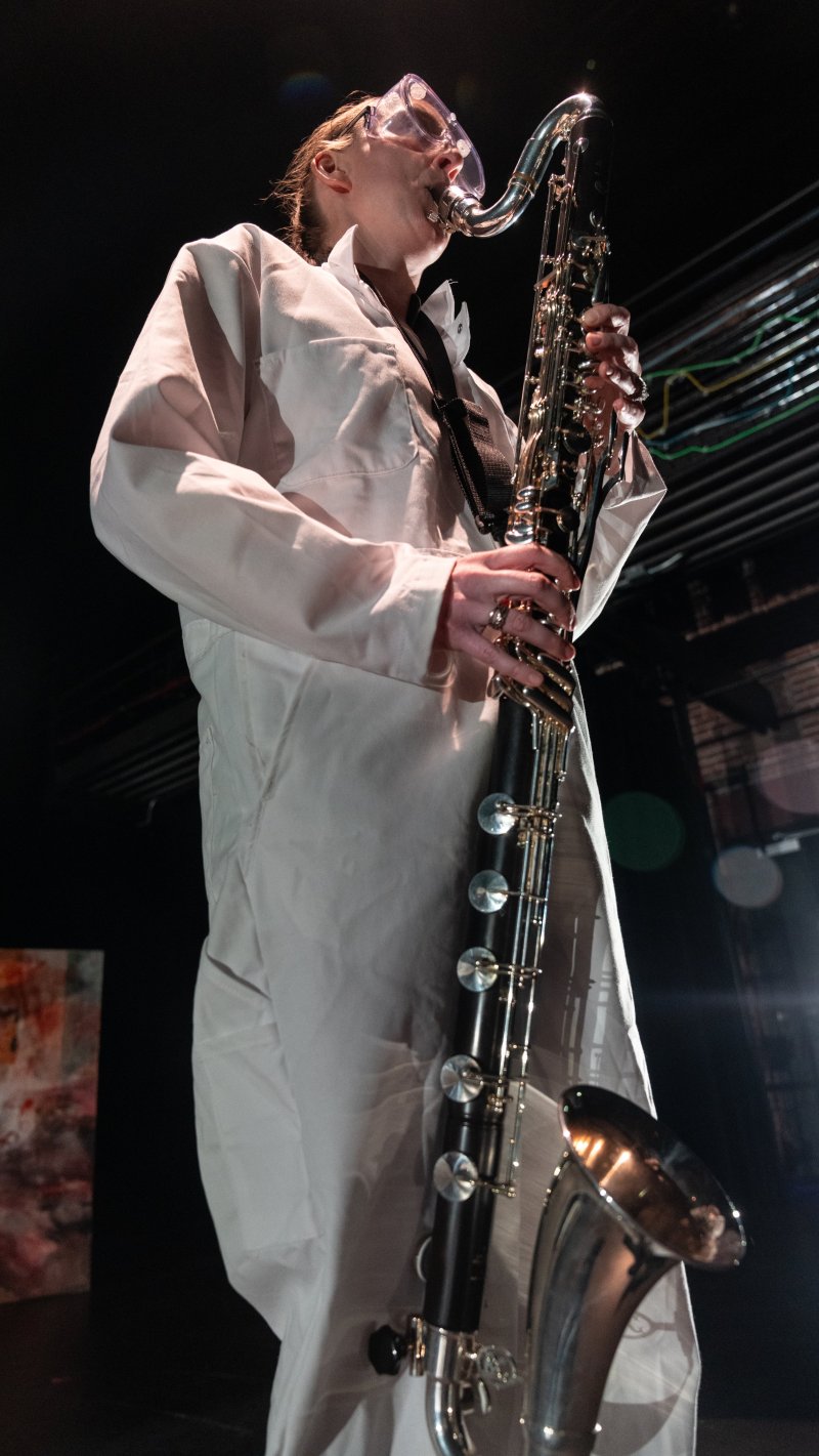 A person in a white lab coat plays a large, dark bass clarinet, with bright stage lights reflecting off the instrument's keys.