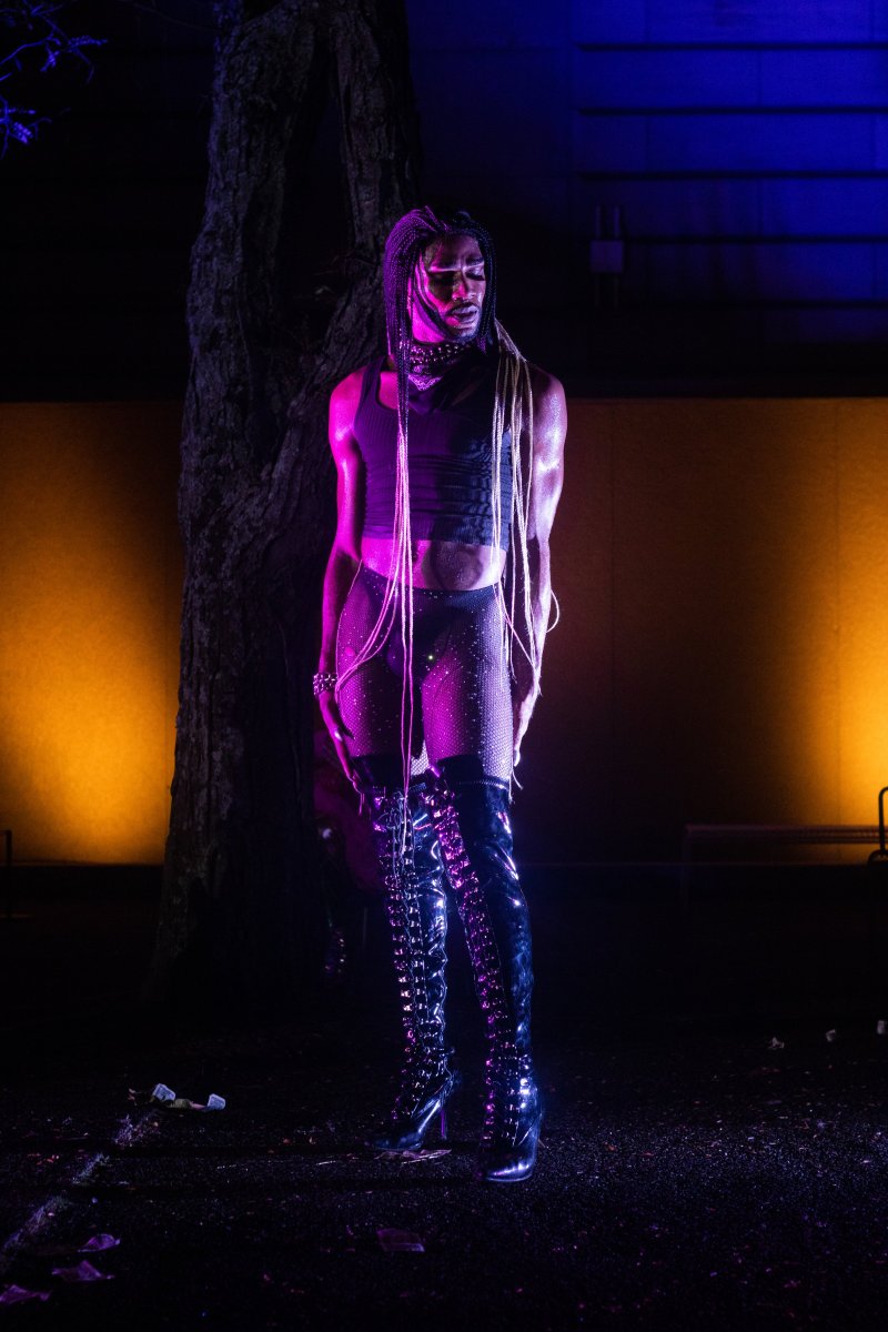 Full-length shot of a person with long braids wearing a black tank top, fishnet bottoms, and shiny black thigh-high boots, standing in front of a tree, lit with pink and blue light.
