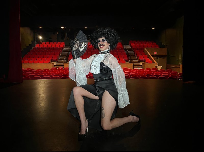 Drag performer in black dress, white ruffled shirt, and black wig, kneeling on stage with a "Slay" fan.