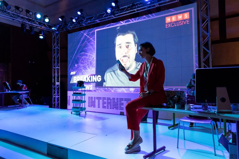 A stage bathed in blue light features a woman in a red suit seated on a desk, holding a phone. Behind her, a large screen displays a man's face and "BREAKING NEWS" graphics. Stage lighting and other figures are visible.