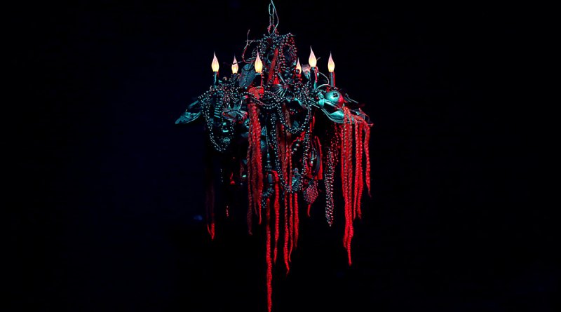 Dark, ornate chandelier with glowing orange candles and long, red, braided strands hanging on a black background.