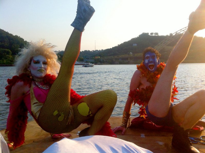 Two performers in drag and body paint pose on a boat with legs raised, river and hills in background at sunset.