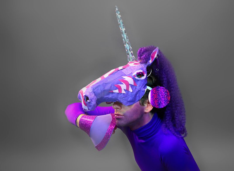 Person in purple bodysuit and unicorn mask with glittery horn, megaphone, and headphones against a gray background.