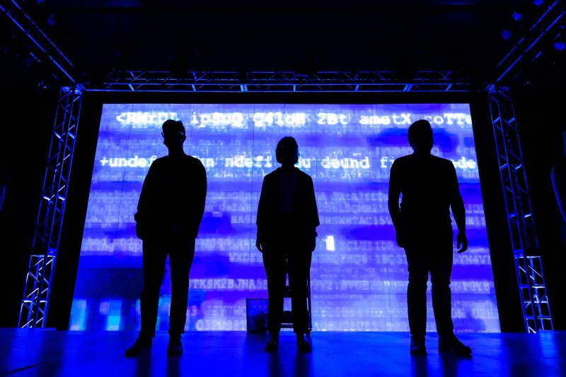 Silhouetted figures of three people stand against a bright blue screen displaying white code. Stage lighting and structural supports are visible.