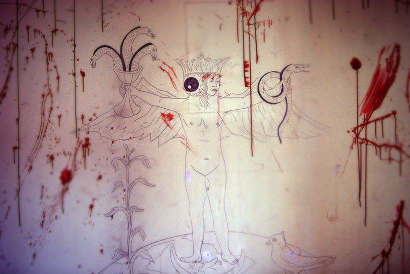 Sketch of a crowned winged figure with snakes, a plant, and a bird, splattered with red paint on a textured wall.