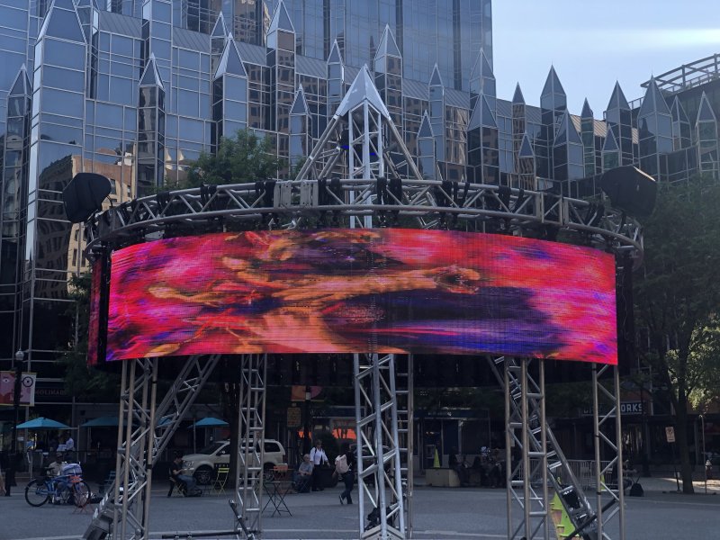 Outdoor video installation featuring a large, curved LED screen displaying abstract patterns in vibrant reds, purples, and oranges. The screen is supported by a silver metal truss structure. A modern glass building is in the background.