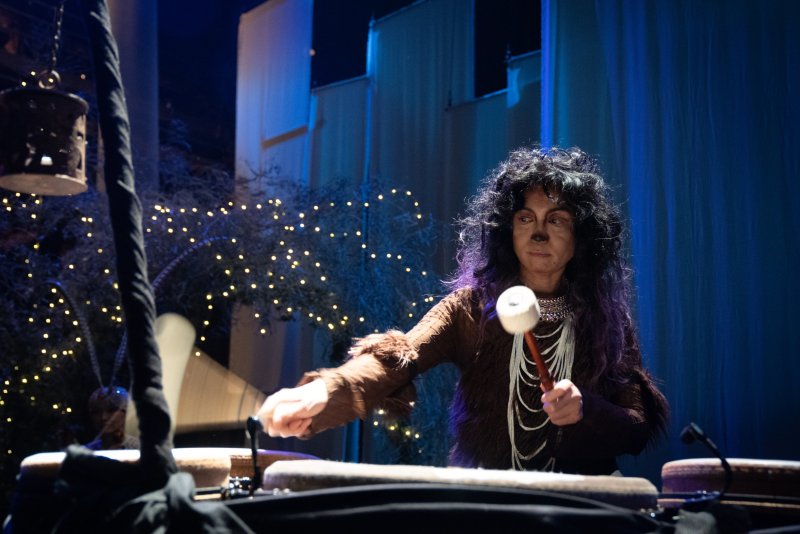 Person in brown furry costume with dark wig and painted nose plays drums with mallets, surrounded by string lights and blue fabric.