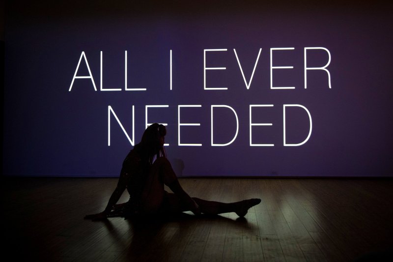 Neon text "ALL I EVER NEEDED" glows on a dark purple wall behind a silhouetted figure sitting on a polished wooden floor.