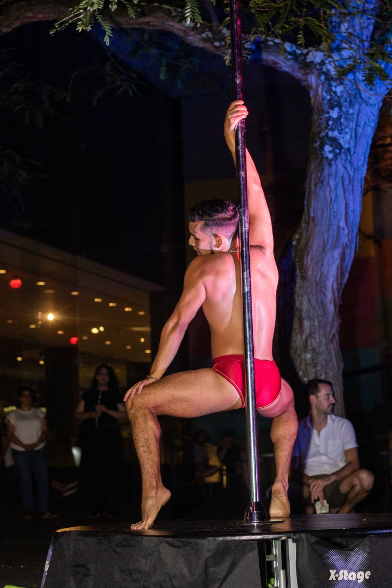 A shirtless man in red briefs performs on a silver pole. The scene is at night with a tree behind him lit with blue light. People are watching in the background.