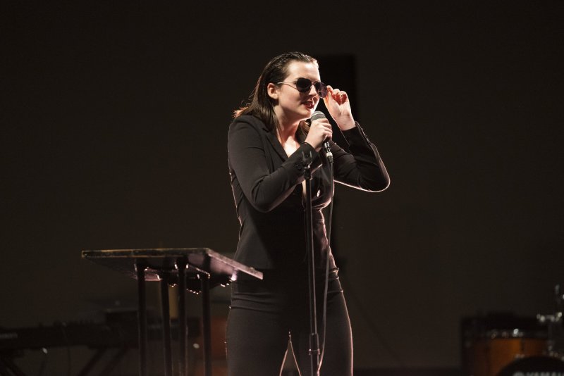 Dark stage view of a performer in black suit and sunglasses singing into a microphone.