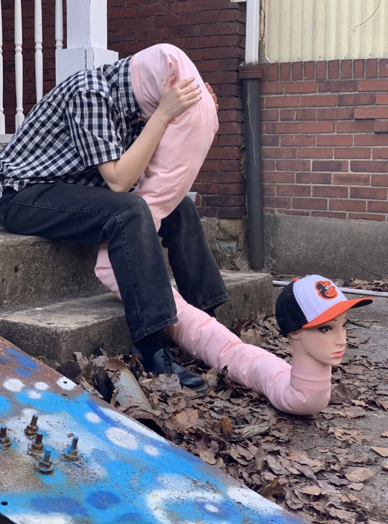 Figure seated on steps, head obscured by a pink pillow. A pink tube extends from the figure, ending in a mannequin head wearing a baseball cap. Debris and leaves surround the base.