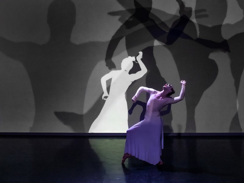 Dancer in a long lavender dress poses on stage. Behind, a wall displays layered black and white silhouettes of figures in similar poses. Stage is dark with a spotlight on the dancer.