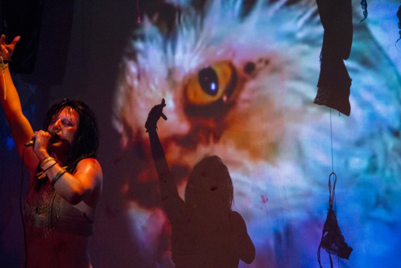 Performance art featuring a singer with a microphone, silhouetted figure, and a large projected image of an owl eye in blue, orange, and white. Dark fabric hangs in the foreground.