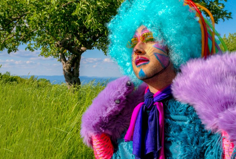 Drag performer in a bright blue wig, purple fur coat, and colorful face paint, outdoors in a grassy field.