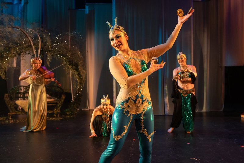 Woman in a sparkly teal and gold costume with a headdress, dancing and holding cymbals, with three other performers in the background.