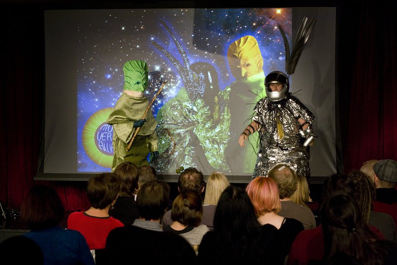 Two performers in alien costumes on stage, one green, one silver, in front of a cosmic projection.