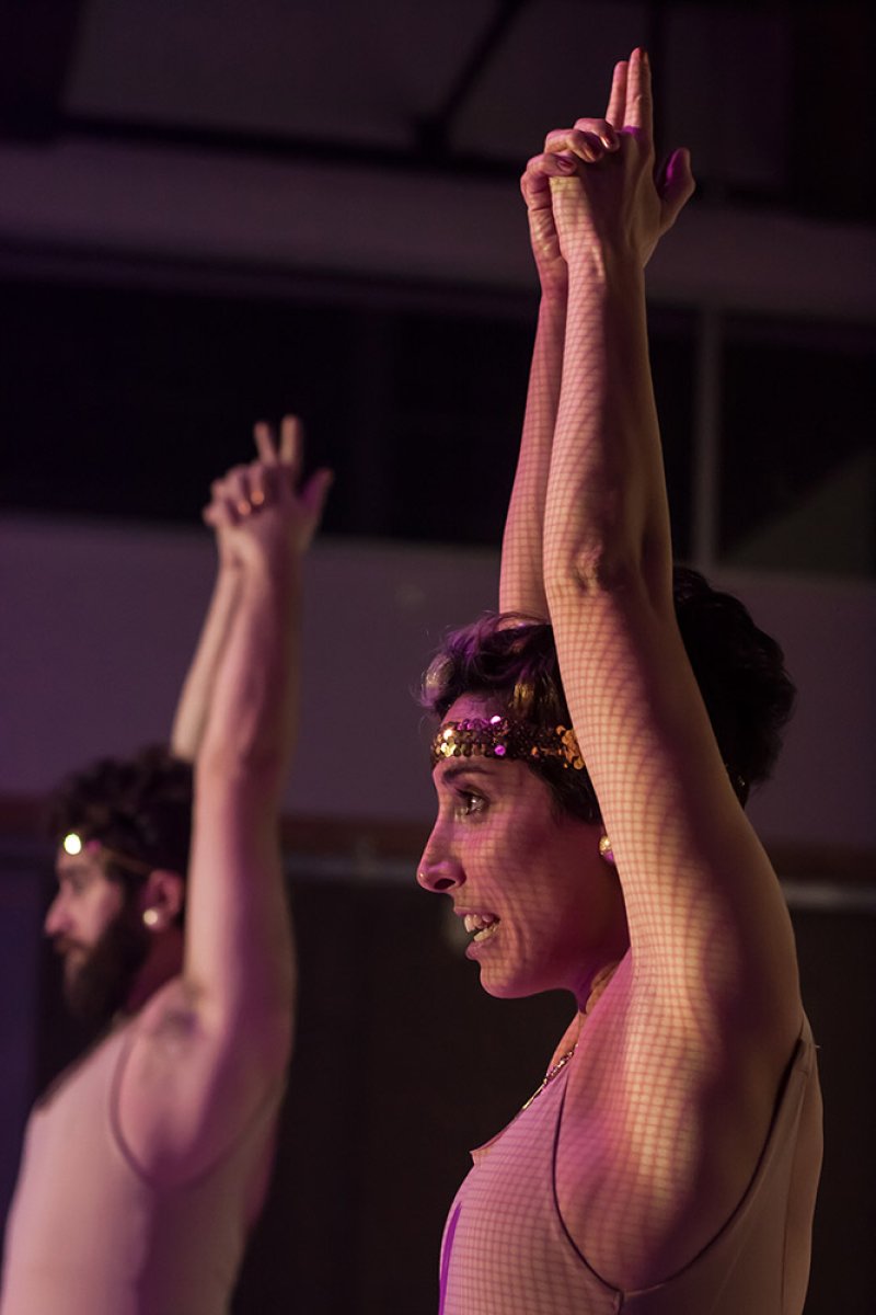 Two dancers, a woman and a man, with arms raised, lit by purple light. Woman wears a gold headband.