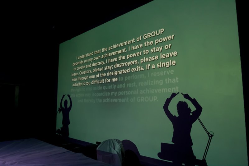 Green projection of text and two dark silhouettes of figures with raised arms on a wall.