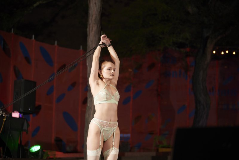 Performance art photograph: A fair-skinned person in lingerie is tied to a tree with black ropes. Red backdrop with blue ovals.