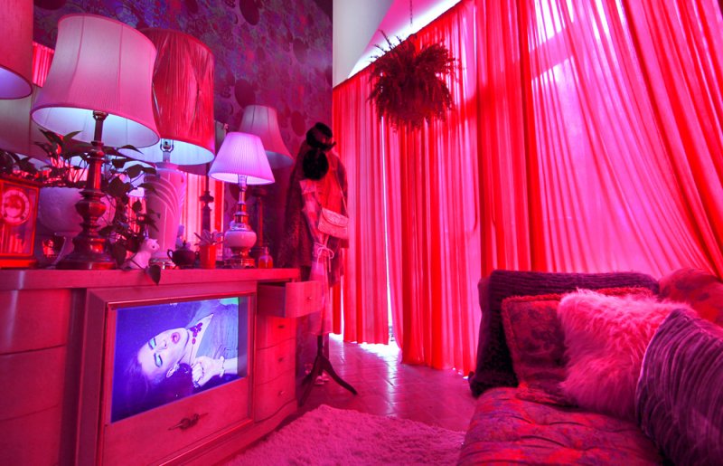 Room bathed in red light, with a dresser, lamps, a mannequin, and a bed. A screen on the dresser shows a woman's face.
