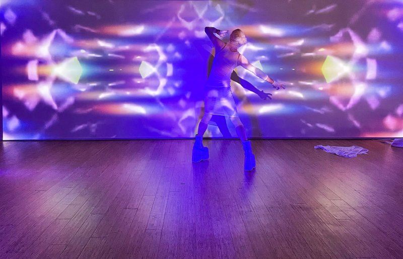 Performer in white, platform boots, striking a pose against a wall projected with vibrant, symmetrical light patterns in purple, blue, and yellow.