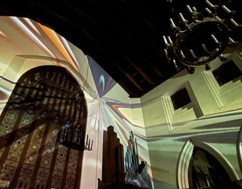 Light projections in warm and cool tones swirl across a church interior with stained glass, organ pipes, and a chandelier.