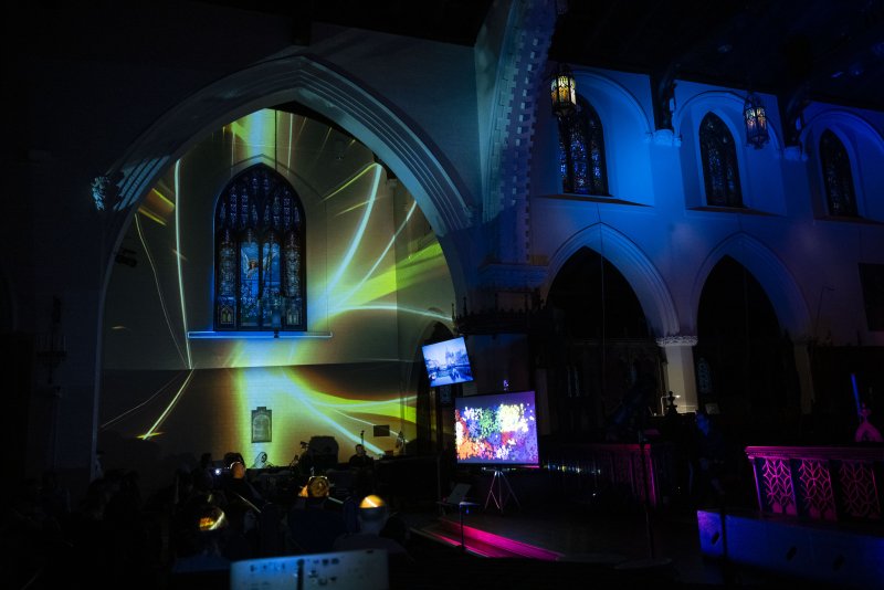 Scott N Andrew, Vox Humana. Dark church interior with projected neon green and yellow light streaks on a stained-glass window and wall.