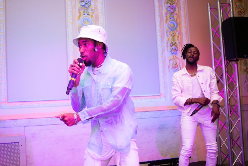 Two performers in white clothing stand on stage under pink and purple lights. The lead singer wears a sheer, light blue button-down shirt and a white bucket hat, holding a microphone. A second performer stands slightly behind, also holding a micropho