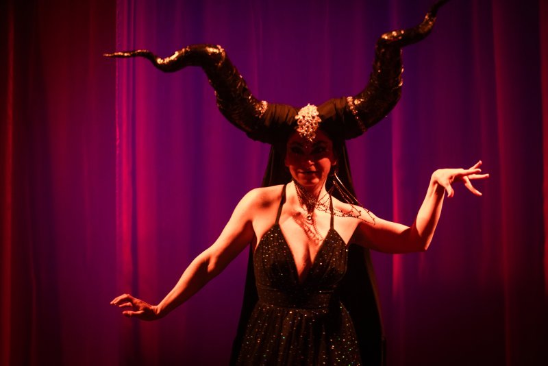 Woman with large black horns, dark dress, and elaborate makeup, posing against a purple-red curtain.