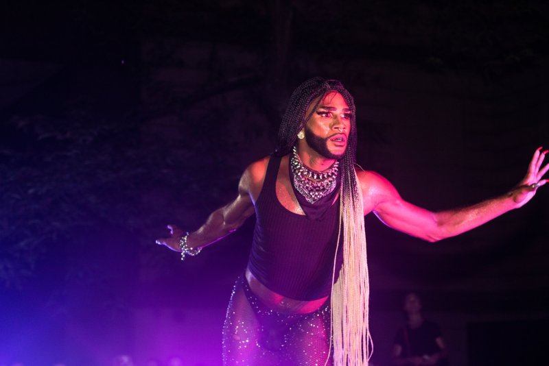 Performer in black tank top and sparkly mesh pants under pink and purple lights. Braided hair, statement necklace, and long red nails. Arms outstretched.