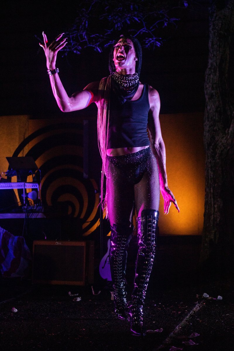 Performer in black tank top, sequined shorts, and thigh-high boots stands on stage with arms raised, lit in pink and purple. Braided hair, studded collar, and dark background with musical equipment visible.