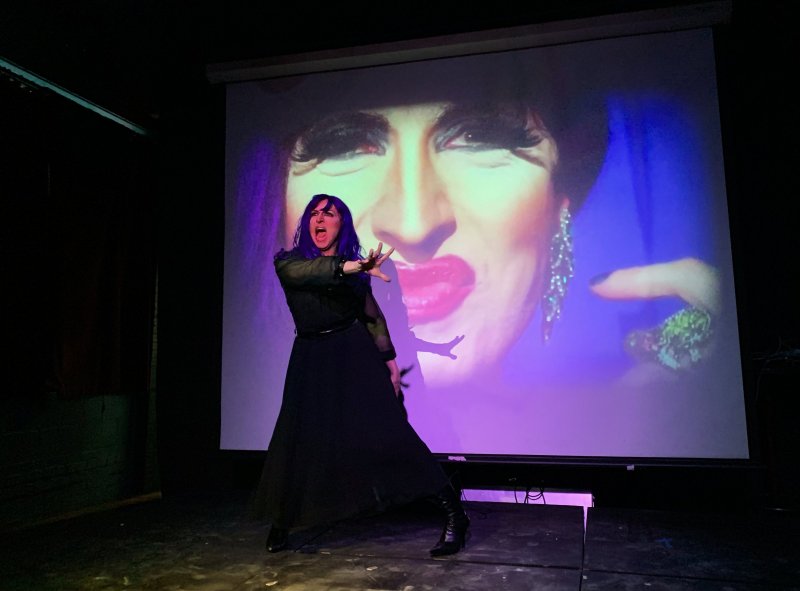Performance art piece: A woman in a black dress and boots stands on stage with arm outstretched, mouth open, against a projection of a face with red lips and a large ring. Predominant colors are black, purple, and red.