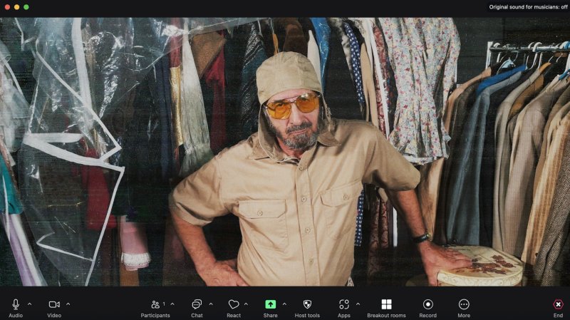 Man in tan hat, shirt, and yellow sunglasses, leaning on a stool in a cluttered room with clothes racks and plastic sheeting.
