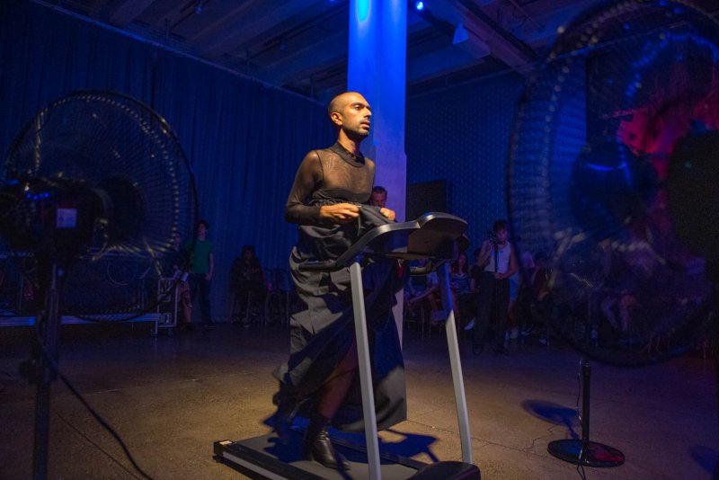 Performance art piece with a bald person in black clothing walking on a treadmill, flanked by large fans, bathed in blue light, with an audience in the background.