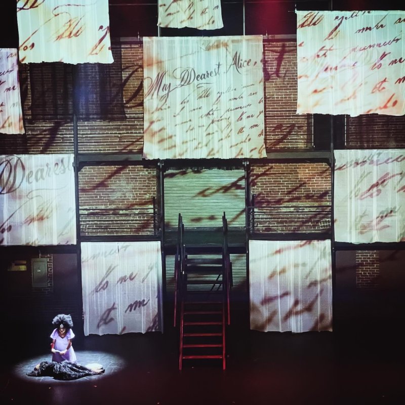 Stage set with brick walls, hanging fabric panels printed with script, red stairs, and two figures in a spotlight.