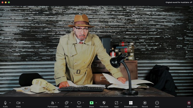 Man in fedora and trench coat leans over a messy desk, looking at papers in a video call interface.
