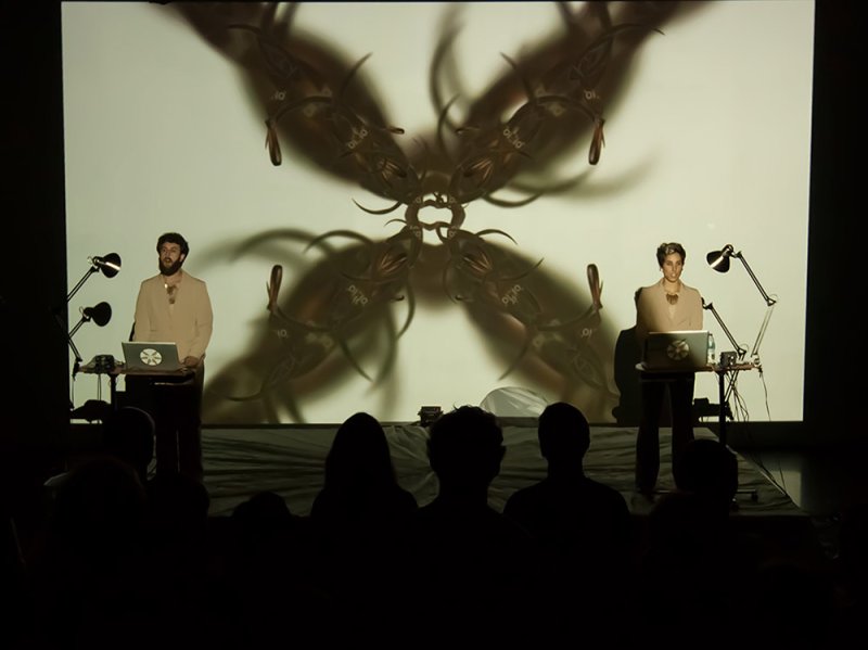 Two performers in light suits at laptops on a stage, with a large, symmetrical, dark brown abstract projection behind them.