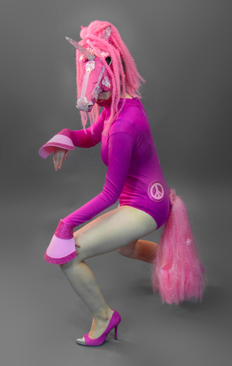 Person in a pink unicorn mask, wig, leotard with a peace sign, and heels, crouching against a gray background.