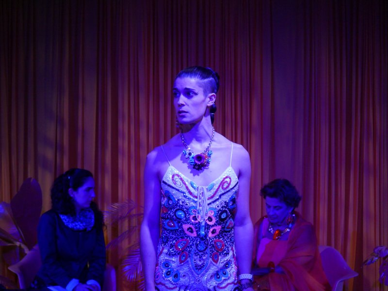 Woman with shaved sides in a colorful beaded dress, wearing a statement necklace, looking right; two women sit behind her.