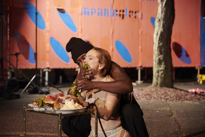 Performance art featuring two figures eating from a table laden with food. One figure feeds the other against a backdrop of orange panels with blue oval shapes.
