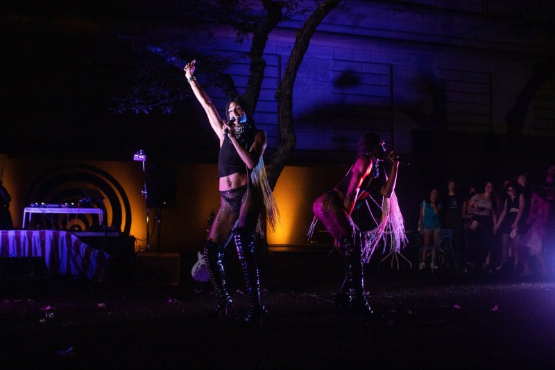 Two figures perform on an outdoor stage with purple and orange lighting. They wear revealing outfits and tall boots. A crowd watches in the background. DJ equipment is visible on the left.